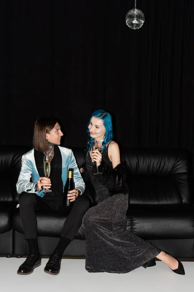 Amigos queer elegantes segurando copos de champanhe enquanto celebra o Natal no sofá de couro no fundo preto — Fotografia de Stock
