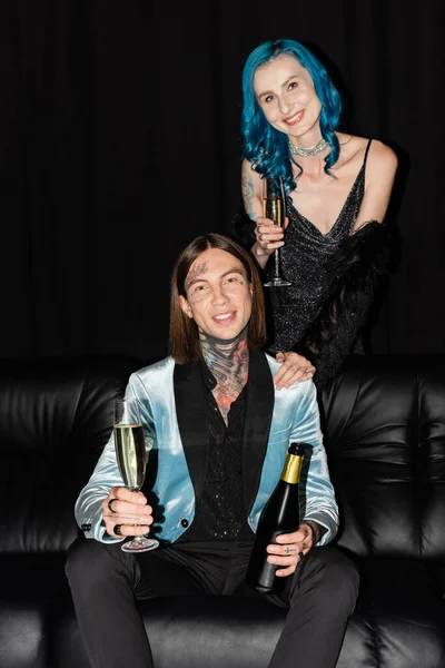 Glückliche und elegante nichtbinäre Freunde mit Champagner, die auf schwarzem Hintergrund in die Kamera lächeln — Stockfoto