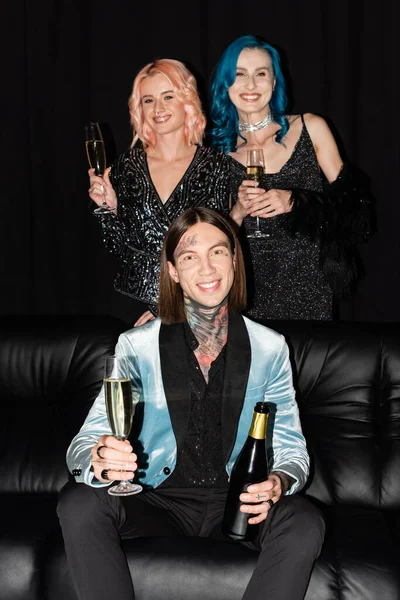 Queere Freunde mit Champagnergläsern in der Hand und lächelnd in der Nähe einer nichtbinären Person, die auf einem Sofa auf schwarzem Hintergrund sitzt — Stockfoto