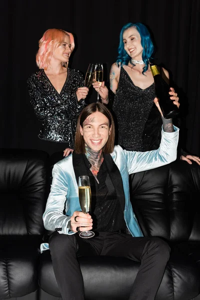 Homme queer tatoué avec bouteille de champagne près d'amis non binaires cliquetis verres sur fond noir — Photo de stock