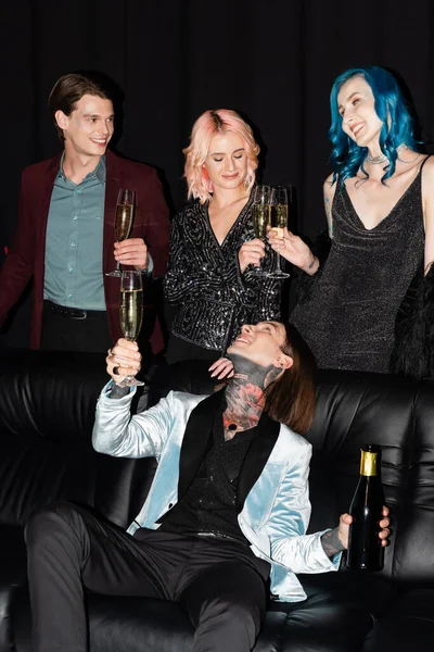 Cheerful tattooed man sitting on couch and looking at elegant nonbinary friends with champagne glasses on black background — Stock Photo