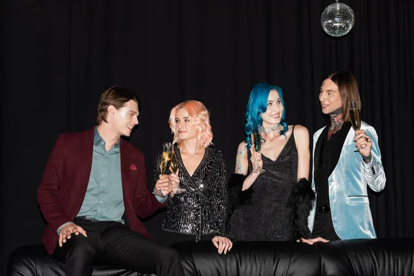 Lächelnde queere Freunde mit Champagnergläsern, die sich während einer Neujahrsparty in der Nähe einer Ledercouch auf schwarzem Hintergrund unterhalten — Stockfoto