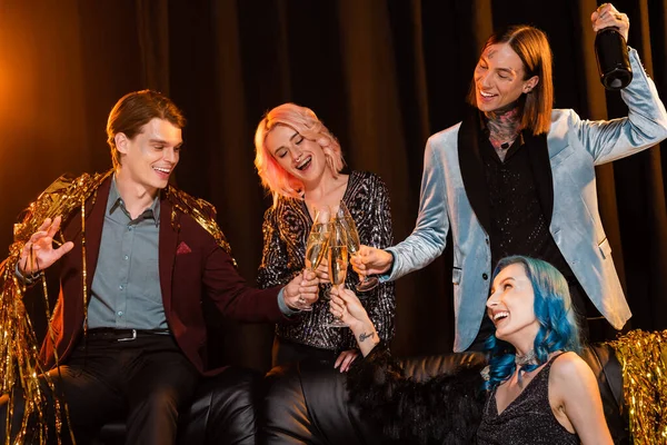 Excited queer friends clinking champagne glasses near golden tinsel on dark background with lighting — Stock Photo