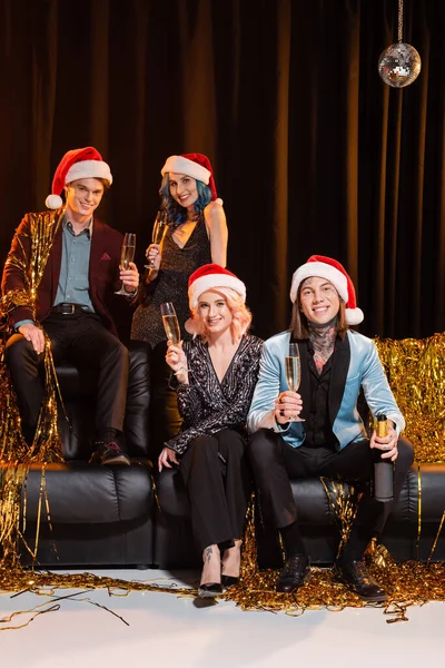Alegre queer amigos em santa chapéus segurando champanhe e celebrando o Natal perto de ouropel no sofá de couro no fundo escuro — Fotografia de Stock