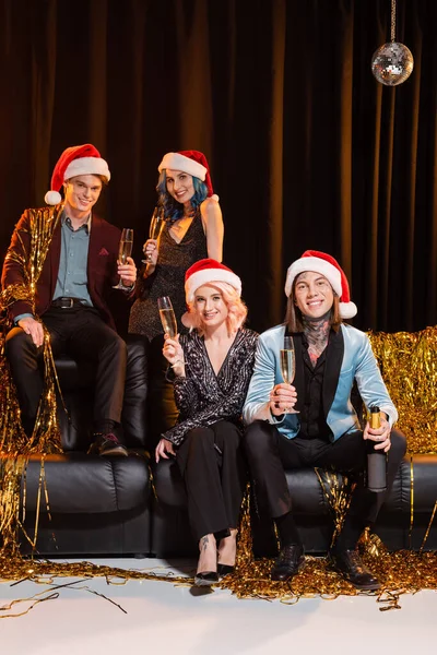 Joyeux amis queer chapeaux Santa tenant champagne et célébrant Noël près de tinsel sur canapé en cuir sur fond sombre — Photo de stock