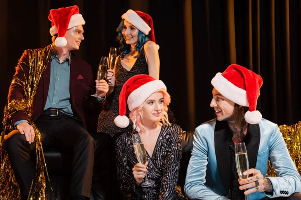 Smiling nonbinary people in santa hats holding champagne and talking during christmas celebration on dark background — Stock Photo