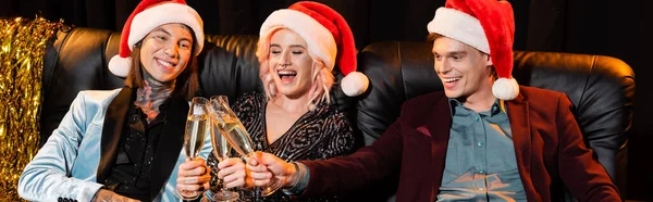 Cheerful nonbinary people in santa hats celebrating christmas and clinking champagne glasses of dark background, banner — Stock Photo