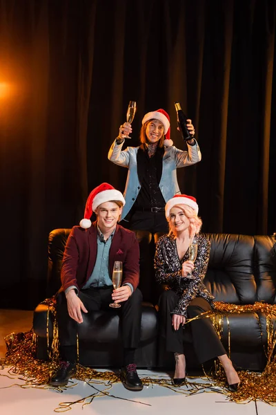 Aufgeregte queere Person, die Champagner in der Nähe nichtbinärer Freunde hält, die auf einer Couch in Weihnachtsmänteln auf dunklem Hintergrund mit Beleuchtung sitzen — Stockfoto