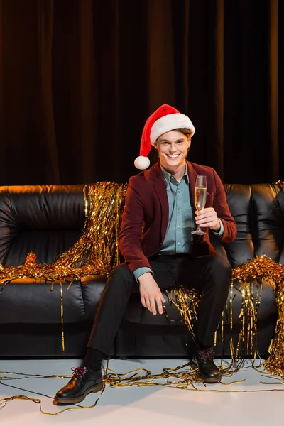 Homem feliz em santa chapéu sentado no sofá de couro com copo de champanhe e olhando para a câmera no fundo escuro — Fotografia de Stock