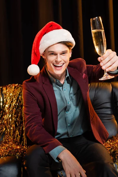 Щасливий чоловік в капелюсі Санта тости з шампанським, сидячи біля золотої мішури на темному фоні — стокове фото
