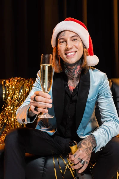 Tattooed queer person in santa hat holding champagne and smiling at camera on dark background — Stock Photo