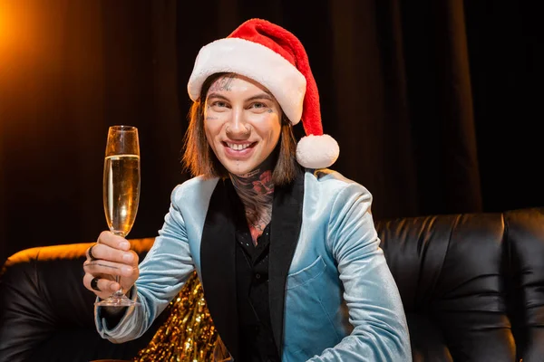 Tatuato persona non binaria in cappello di Babbo Natale in possesso di champagne e guardando la fotocamera su sfondo scuro con illuminazione — Foto stock