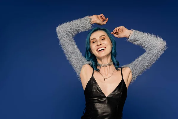 Cheerful woman in black leather dress and fluffy sleeves dancing with raised hands isolated on blue — Stock Photo