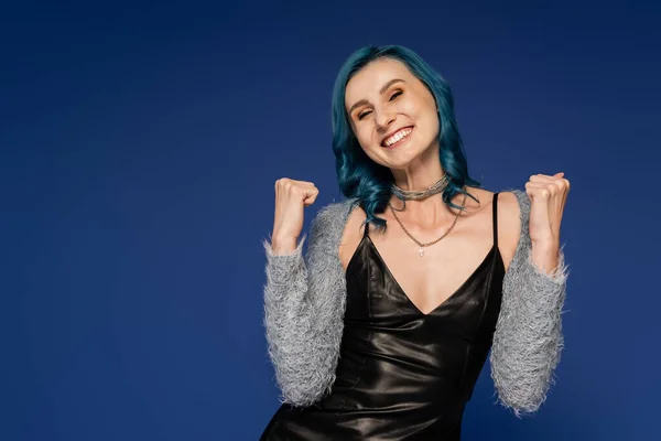 Excited woman in black leather jacket and faux fur sleeves showing win gesture isolated on blue — Stock Photo