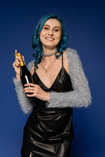 Elegante donna in pelle nera vestito in possesso di bottiglia di champagne e sorridente alla fotocamera isolata su blu — Foto stock