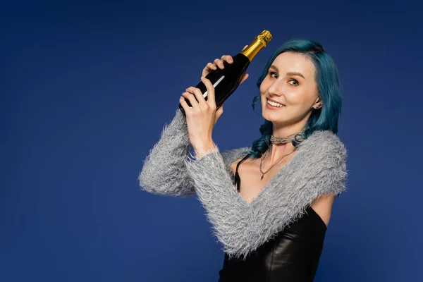 Радостная и модная женщина с бутылкой шампанского, смотрящая на камеру, изолированную на голубом — стоковое фото