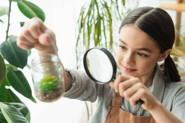 Menina sorrindo segurando lupa e frasco borrado com suculento em casa — Fotografia de Stock