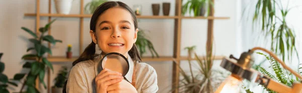 Cheerful girl holding magnifying glass and looking at camera near blurred lamp at home, banner — Stock Photo