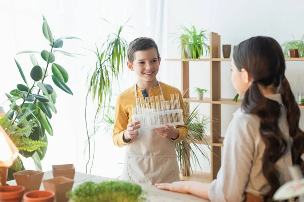 Sonriente niño sosteniendo tubos de ensayo cerca borrosa amigo y plantas en casa - foto de stock