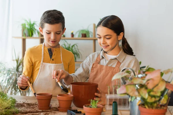 Souriante fille versant le sol dans le pot de fleurs près de l'ami et des plantes à la maison — Photo de stock