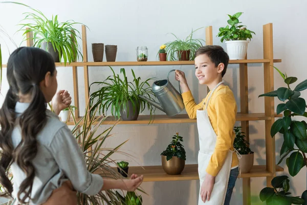 Sonriente niño riego planta cerca borrosa amigo en casa - foto de stock