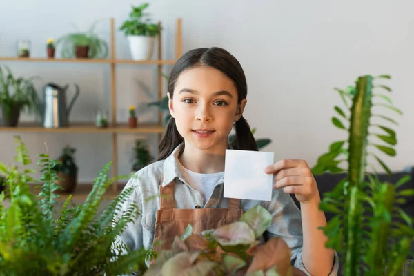 Preteen fille dans tablier tenant note collante près de plante floue à la maison — Photo de stock