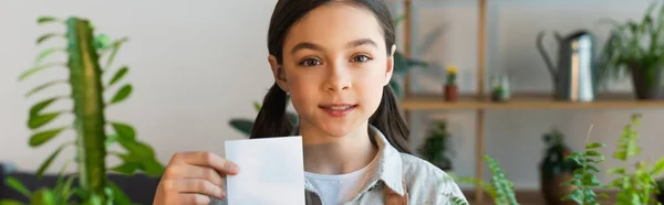 Sonriente niño sosteniendo nota pegajosa cerca de las plantas borrosas en casa, pancarta - foto de stock