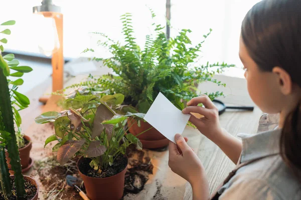 Kind hält klebrigen Zettel in der Nähe von Pflanzen und Erde auf dem heimischen Tisch — Stockfoto