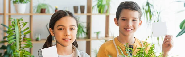 Enfants souriants tenant des notes collantes près des plantes à la maison, bannière — Photo de stock