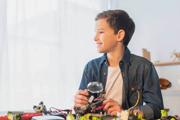 Smiling preteen boy holding magnifying glass near robotics model at home — Stock Photo