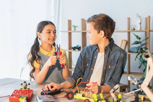 Smiling girl holding pliers near friend making robotic model at home — Stock Photo