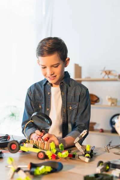 Ragazzo sorridente che tiene lente d'ingrandimento vicino al modello robotico offuscato a casa — Foto stock