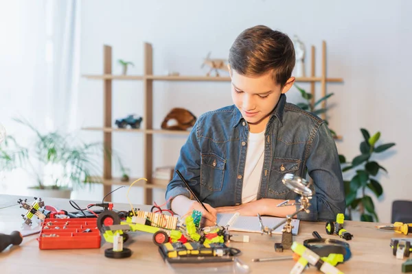 Preteen child writing on notebook near tools and robotic model at home — Stockfoto