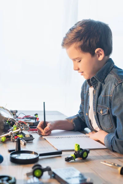Вид сбоку дошкольного мальчика, пишущего на блокноте рядом с увеличительным стеклом и роботизированной моделью дома — стоковое фото