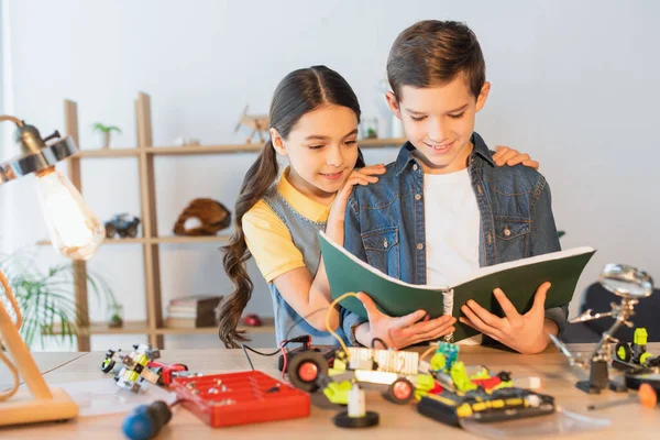 Preteen girl umarmt Freundin mit Notizbuch in der Nähe verschwommenes Robotik-Modell zu Hause — Stockfoto