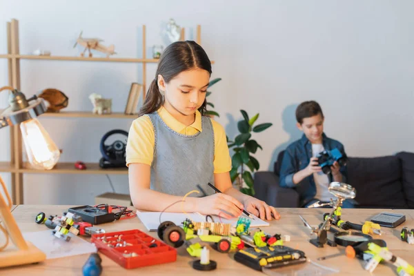 Chica preadolescente escribiendo en un cuaderno cerca de modelo robótico y amigo borroso en casa - foto de stock