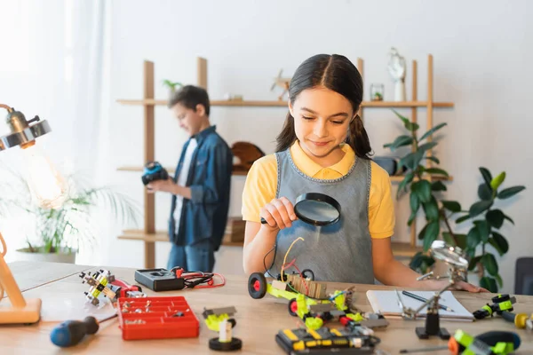 Sorridente bambino preadolescente in possesso di lente d'ingrandimento vicino al modello di robotica a casa — Foto stock