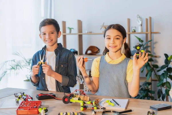 Niños felices sosteniendo herramientas cerca del modelo robótico en la mesa en casa - foto de stock