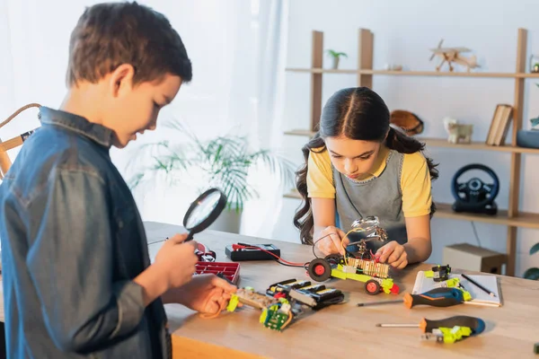 Verschwommener Junge hält Lupe neben Mädchen, das Robotermodell auf dem heimischen Tisch zusammensetzt — Stockfoto