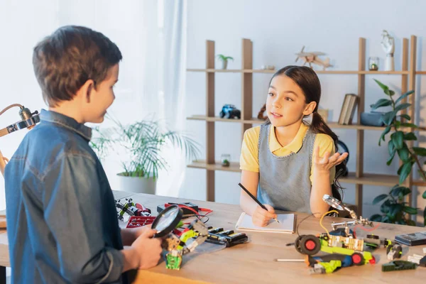 Preteen girl with notebook and pencil talking to friend holding magnifier near parts of robotics model — Stock Photo