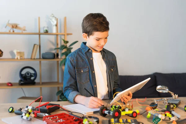 Junge mit digitalem Tablet schreibt in Notizbuch neben Details des Robotermodells auf dem Tisch — Stockfoto