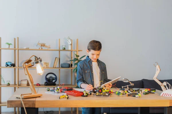 Preteen boy holding digital tablet and writing in notebook near mechanical details of robotics model — Stock Photo