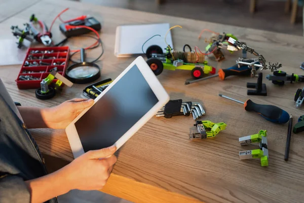 Teilansicht des Kindes, das ein digitales Tablet mit leerem Bildschirm in der Nähe von Werkzeugen und mechanischen Details auf dem Tisch hält — Stockfoto