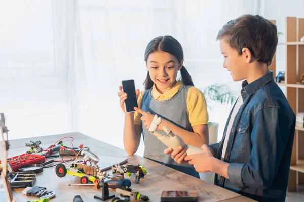 Erstauntes Mädchen zeigt auf Smartphone mit leerem Bildschirm neben Junge, der Roboterhand zu Hause hält — Stockfoto