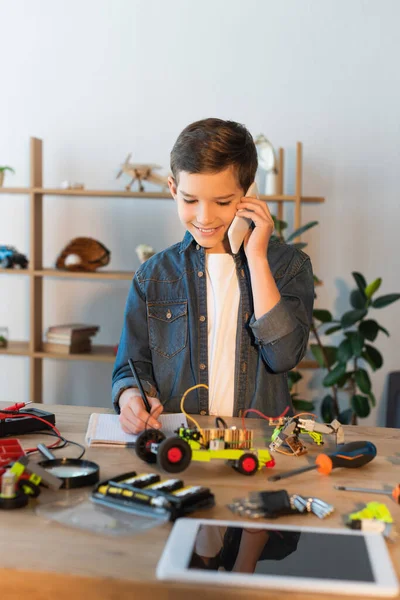 Lächelnder Junge spricht auf Smartphone und schreibt in Notizbuch neben mechanischen Teilen und verschwommenem digitalen Tablet — Stockfoto