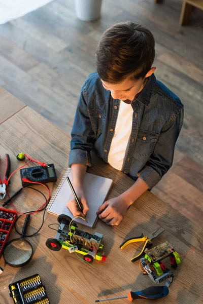 Vista dall'alto del ragazzo che scrive nel quaderno vicino agli strumenti e dettagli del modello di robotica sul tavolo — Foto stock