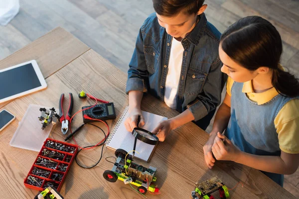 Vista de ángulo alto del niño sosteniendo la lupa cerca del cuaderno y apuntando con lápiz al modelo de robótica - foto de stock