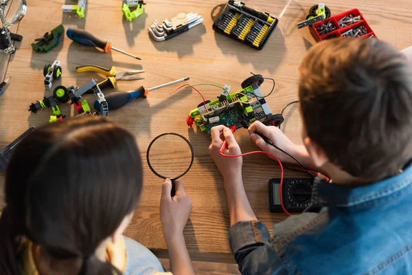 Top view of boy with multimeter and girl with magnifying glass assembling robotics model at home — Stock Photo