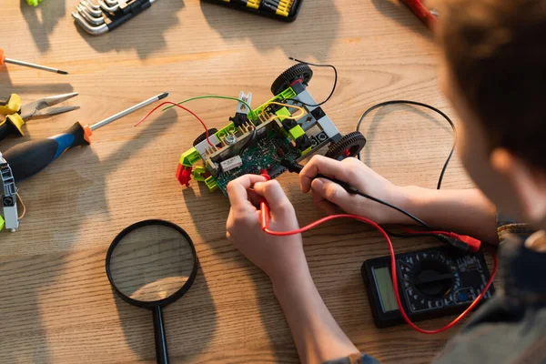 Top view of cropped boy with multimeter measuring voltage of robotics model near magnifier — Stock Photo