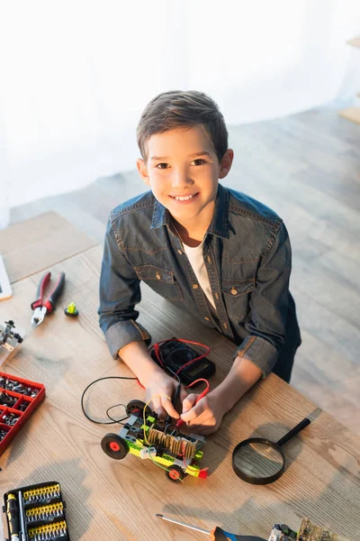 High angle view of smiling boy with multimeter measuring voltage of robotics model near magnifier — Stock Photo